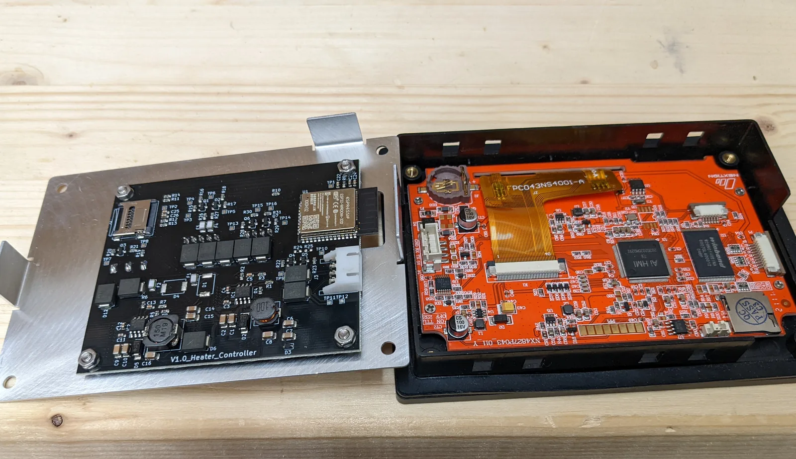 Zwei Leiterplatten nebeneinander auf einer Holzplatte: links eine schwarze Platine mit Funkmodul, Spulen und der Aufschrift V1.0_Heater_Controller, auf ein gekantetes Aluminiumblech geschraubt, rechts eine orangefarbene Displayplatine, über ein Flachbandkabel mit dem Bildschirm verbunden