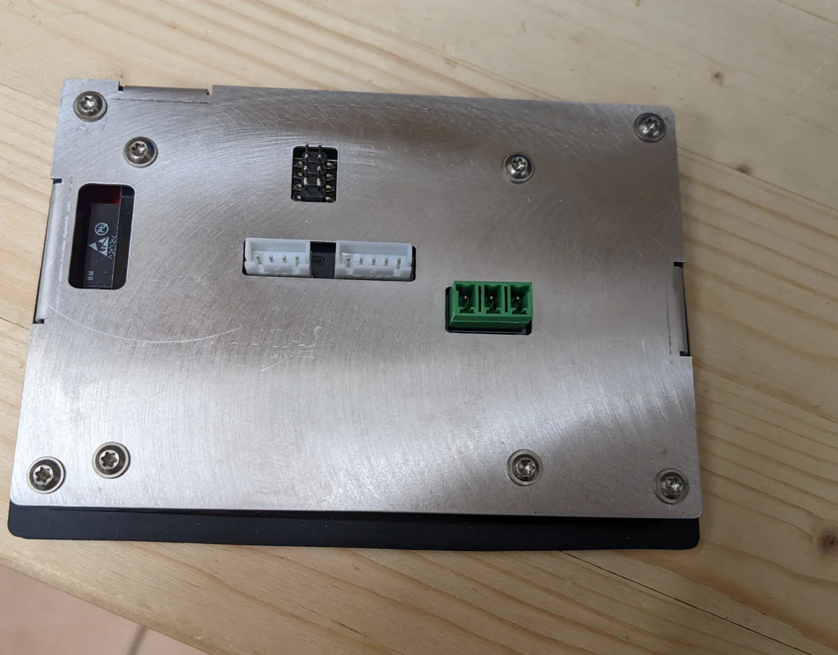 Geschlossenes Bedienteil von hinten: eine gebürstete Aluminiumplatte, mit acht Schrauben am schwarzen Rahmen befestigt, mit Ausschnitten für eine schwarze Stiftleiste, zwei weiße Steckverbinder und eine grüne dreipolige Schraubklemme
