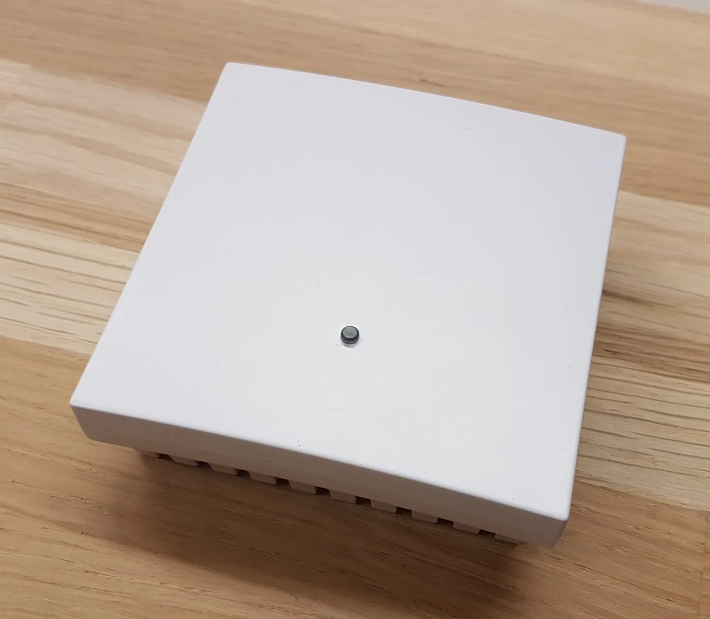 Flacher weißer Wandsensor der GeCoS-Serie auf einer Holzfläche, schräg von oben: geschlossenes quadratisches Gehäuse mit der Messöffnung in der Mitte und Lüftungsschlitzen an der Seite