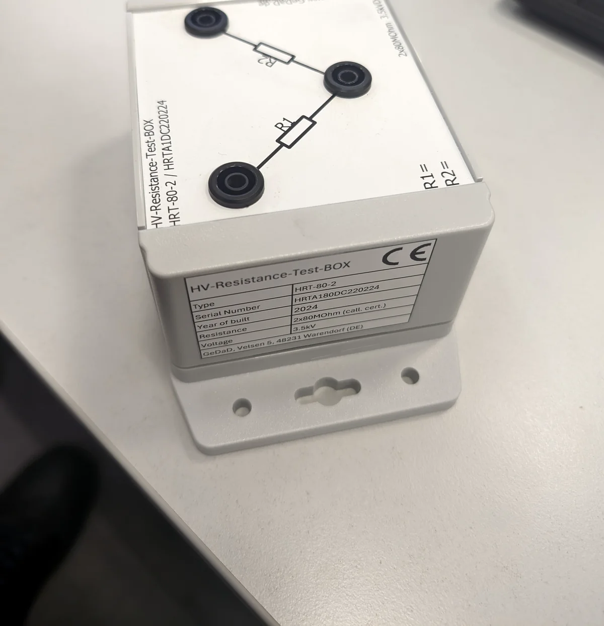Prüfbox HRT-80-2 im weißen Gehäuse mit Wandhalterung, auf der Oberseite drei Hochspannungsbuchsen und das aufgedruckte Schaltbild der beiden Widerstände, auf der Stirnseite das Typenschild von GeDaD mit Typ, Seriennummer, Baujahr 2024, 2 × 80 Megaohm und 3,5 Kilovolt